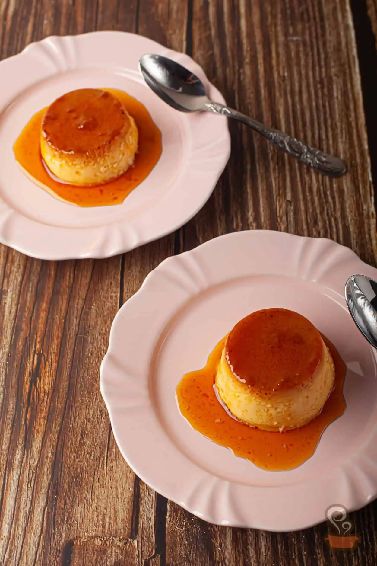 A imagem mostra duas porções de pudim de leite com laranja, servidas em pratos rosa com colheres vintage, sobre superfície de madeira, destacando a textura e brilho da calda.
